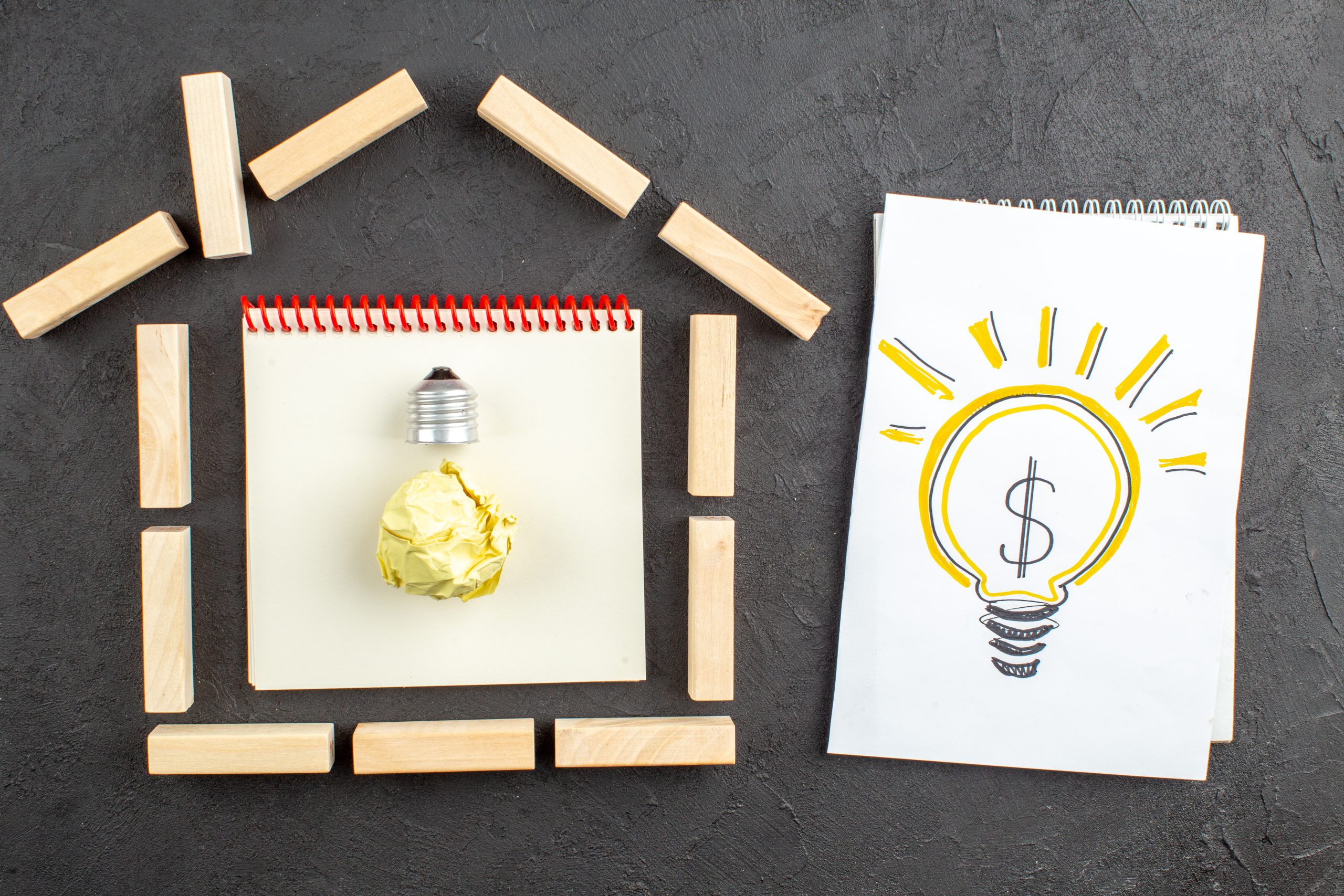 Rysunek przedstawiający żarówkę z symbolem oszczędzania energii na notatniku, ułożonym na drewnianych blokach – symbol alternatywnych inwestycji w oszczędzanie energii