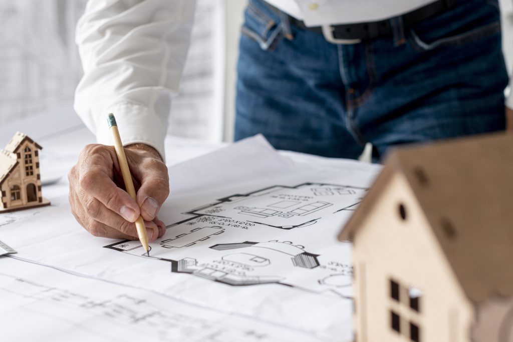Zbliżenie na dłoń architekta nanoszącego poprawki ołówkiem na techniczny plan budynku, obok makieta przedstawiająca nowoczesny dom inwestycyjny.